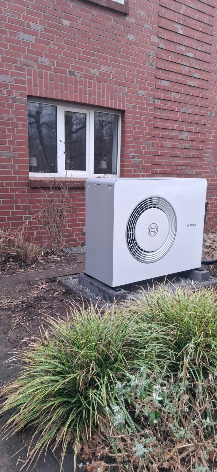 Eine weiße Bosch-Wärmepumpen-Außeneinheit steht auf einem Betonfundament vor einer roten Klinkerhauswand im Garten.